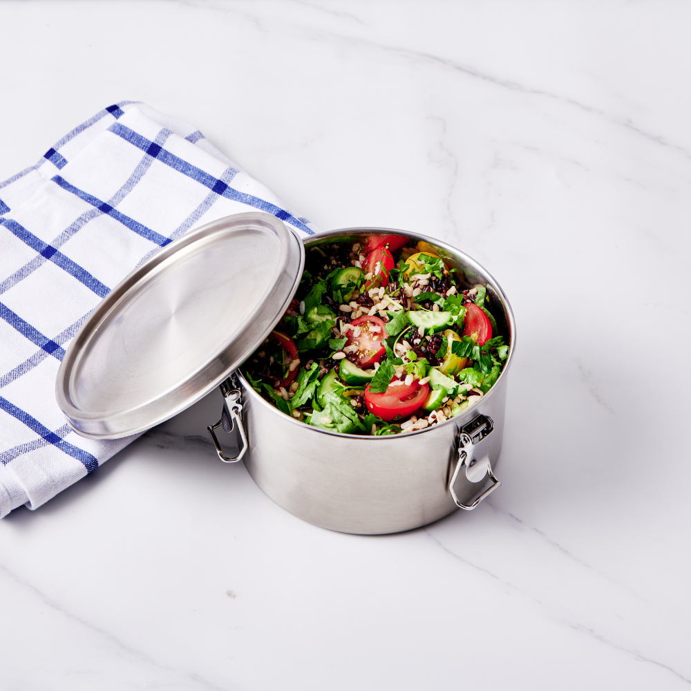 Stainless steel round lunch box with a lid, filled with a colorful salad and placed on a marble surface with a checkered c...