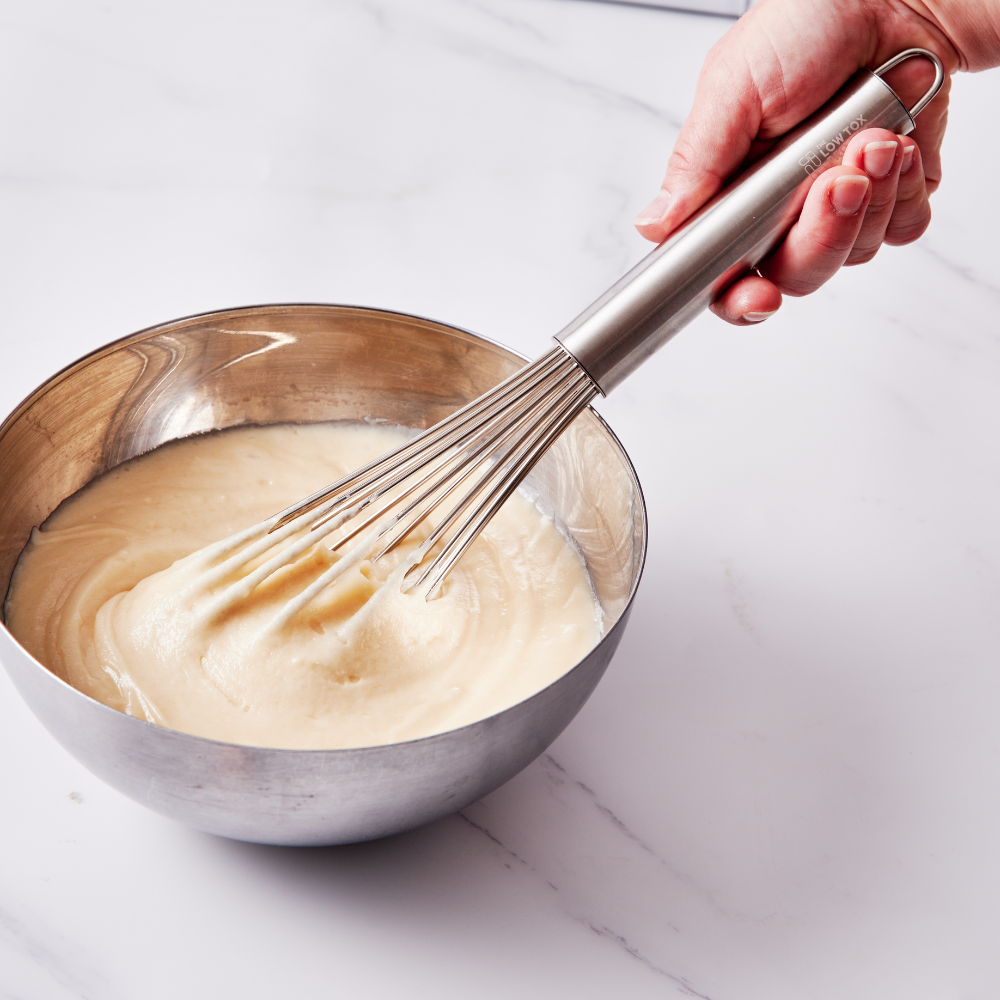 Heavy duty stainless steel whisk resting inside a metal mixing bowl on a neutral background.