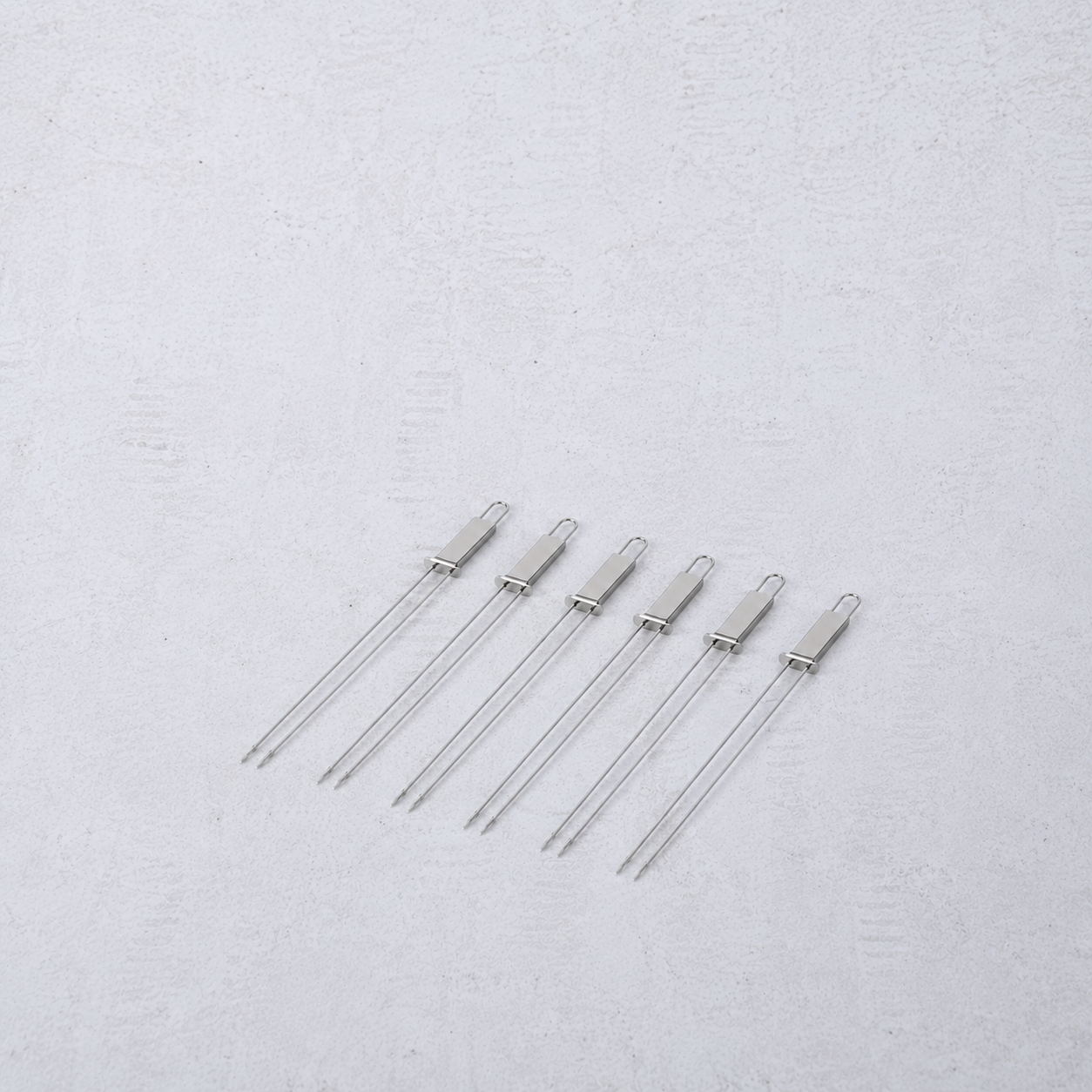 Set of six reusable stainless steel double-prong barbecue skewers with loop handles on a light textured background.